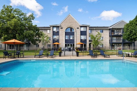 A swimming pool in front of a building with a patio and chairs.