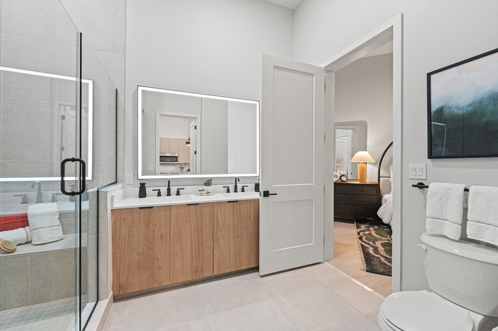 A bathroom with a glass shower door, a white toilet, a sink with a mirror above it, and a rug on the floor.