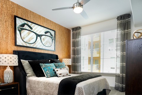 A bedroom with a bed, pillows, a ceiling fan, and a picture of glasses on the wall.