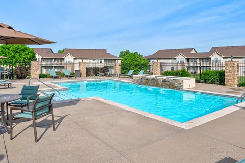 4-Bedroom Apartments in Lawrence, KS - Ironwood Apartments and Townhomes - Swimming Pool with Small Waterfalls, Lounge Chairs, Outdoor Dining Table and Chairs, Umbrellas, and Metal Fence.