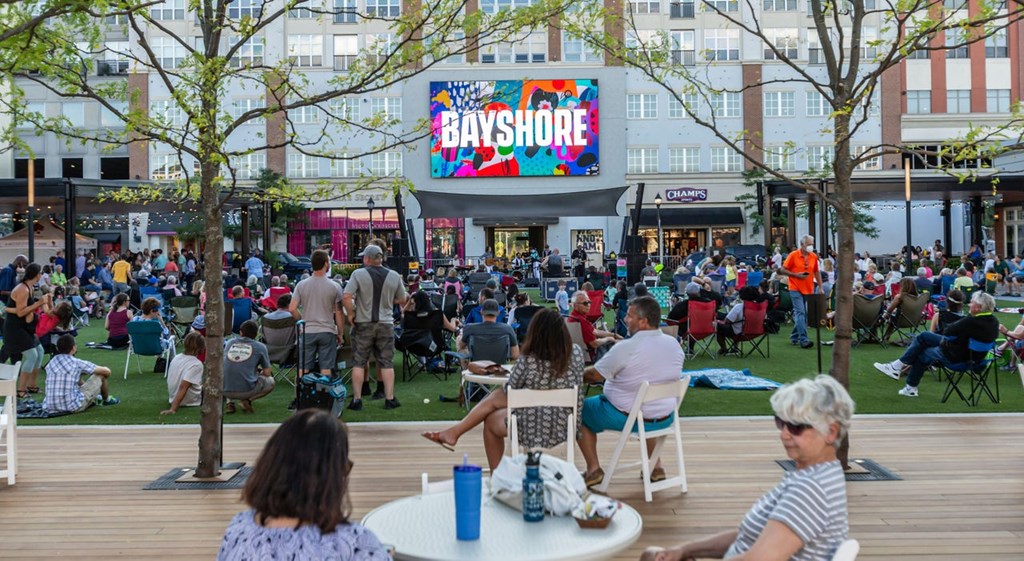 a crowd of people sit on a grassy area in front of a building with a large