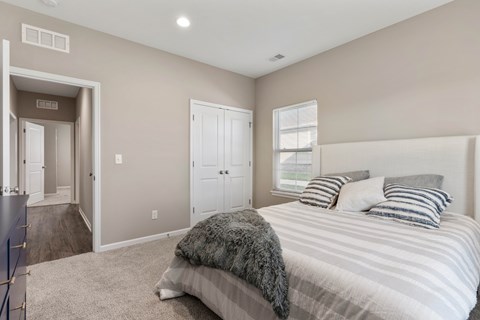 Senior Housing in Belton, MO - Traditions Villas 55+ - Bedroom with Carpet Flooring, High Ceiling, Light Fixtures, Large Bed, Dresser, Window with Blinds, and Doorway.