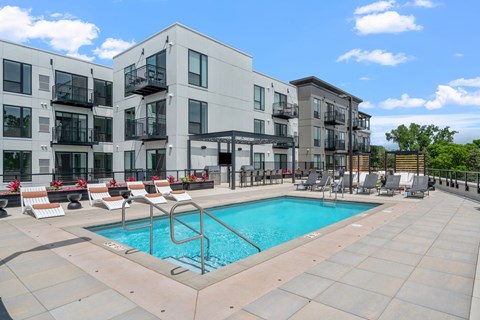 A swimming pool is surrounded by lounge chairs in front of apartment buildings.