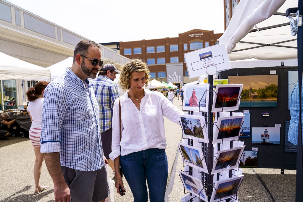 a man and a woman walk past a postcard stand at a farmers market