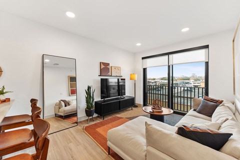 A modern living room with a white sofa, a wooden chair, a television, and a large window with a view of the city.
