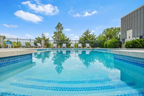 Lawrence, KS, Apartments for Rent - Fairway Flats & The Heights at Fairway Flats - Resort Style Pool With Blue Tile Steps, Lounge Seating, Black Metal Fence, Green Landscaping, And Open Sky.