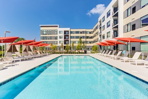 A large swimming pool with red umbrellas and chairs is surrounded by apartment buildings.