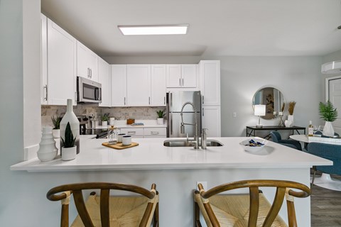 A kitchen with white cabinets and a white countertop with two chairs in front of it.
