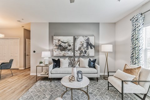 A living room with a grey and white patterned rug, a white sofa, a grey armchair, a coffee table, and a large abstract painting on the wall.