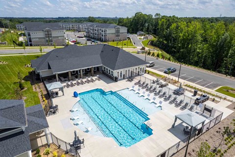A large swimming pool with lounge chairs and a pavilion is surrounded by a parking lot and buildings.