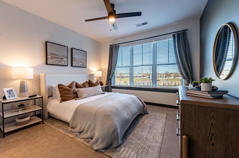 North Indianapolis, IN, Apartments Near Palladium - North End Apartments - Primary Bedroom With Plush Bed, Ceiling Fan, Nightstands, Lamps, Dresser, and Wide Window With Curtains.