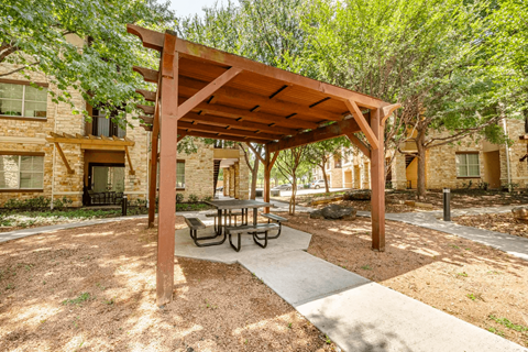 A wooden pergola with a table and chairs is situated in a courtyard surrounded by buildings.