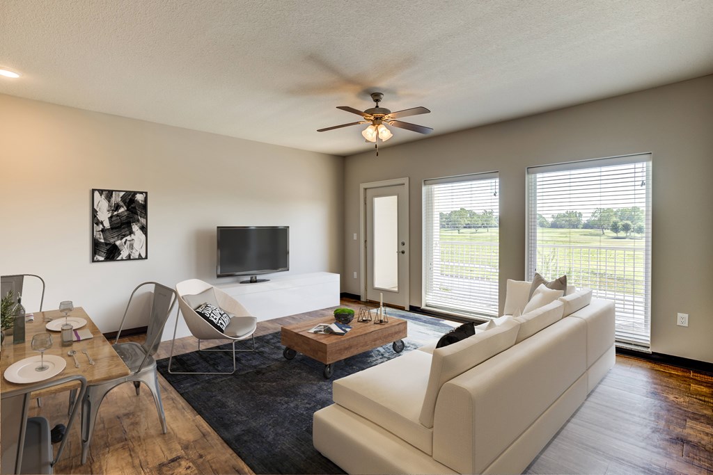Fairway Flats - Spacious Living Area with Large Windows, Wood Flooring, Ceiling Fan, and White Walls