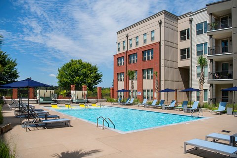 A swimming pool surrounded by lounge chairs and umbrellas in front of a multi-story building.