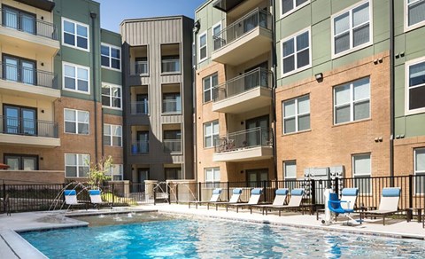 A swimming pool is surrounded by lounge chairs in front of apartment buildings.