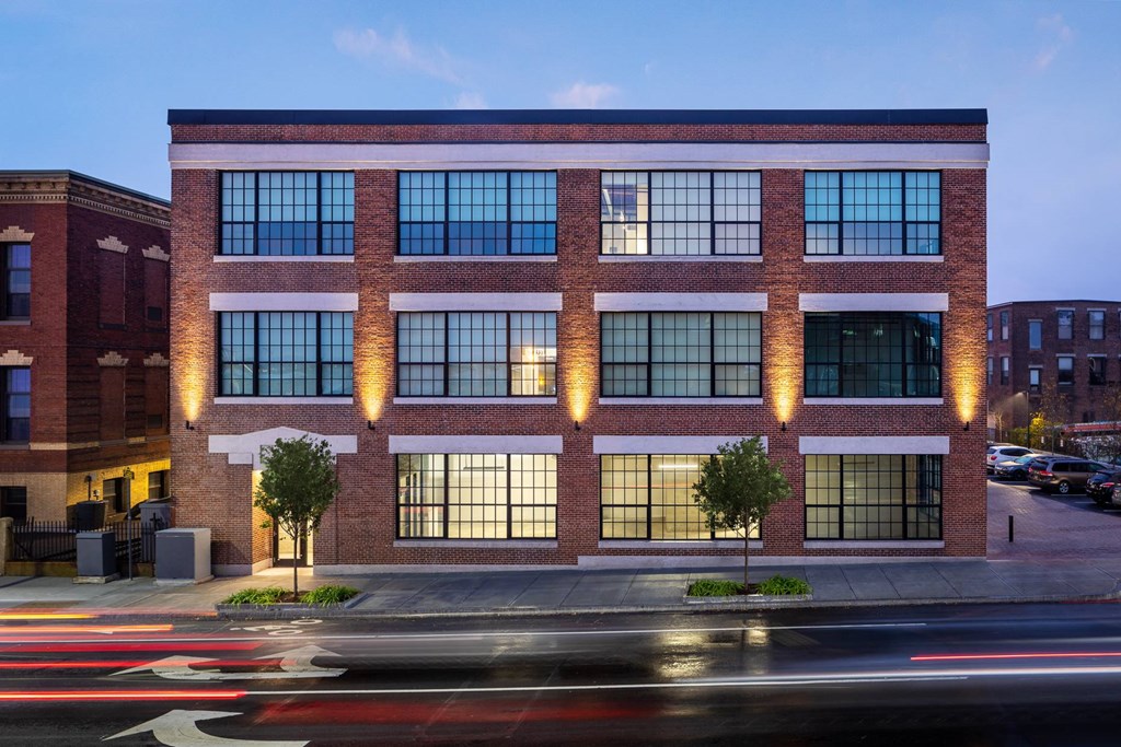 A modern building with a brick facade and large windows is illuminated from within at dusk.