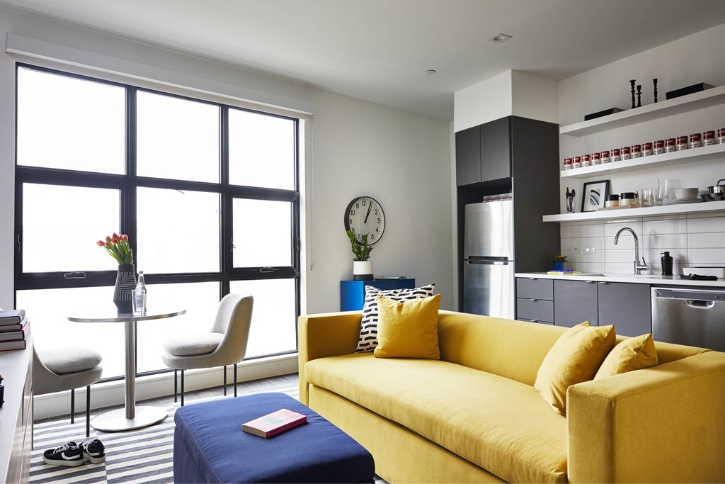A yellow couch with a blue ottoman and a vase with flowers on a table in front of a window.