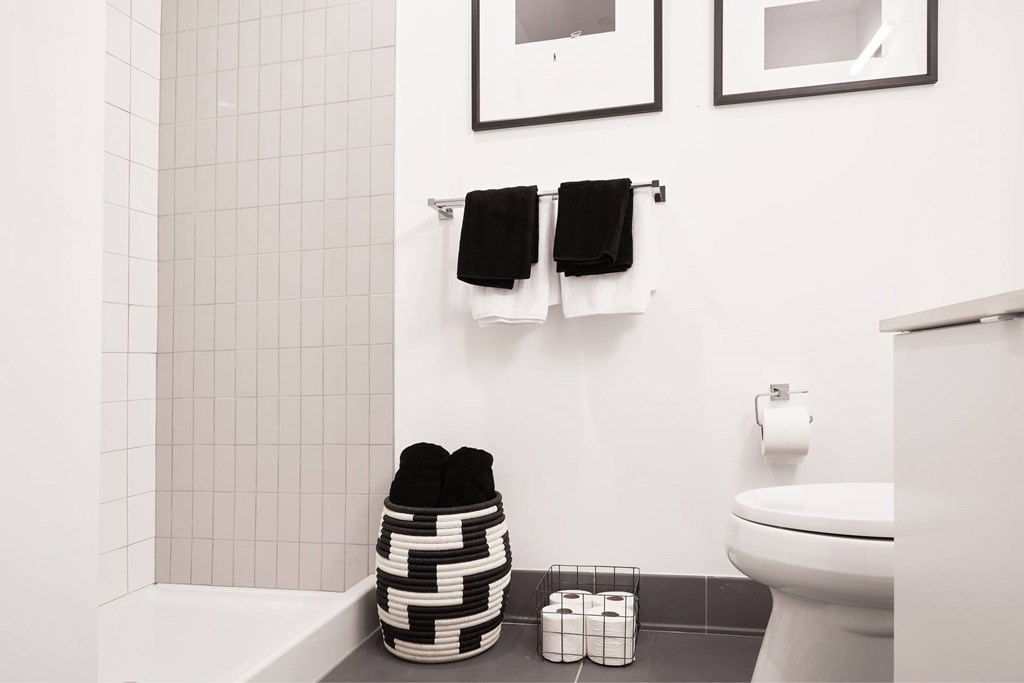 A bathroom with a toilet, a towel rack with two towels, a basket with a black and white bag, and a roll of toilet paper.