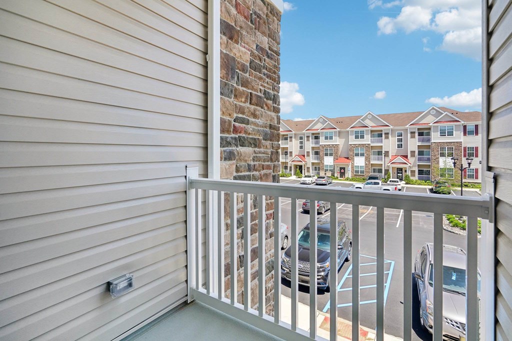 A balcony with a white railing overlooks a parking lot and apartment building.