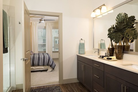 A bathroom with a mirror, sink, and a bedroom with a bed and window.