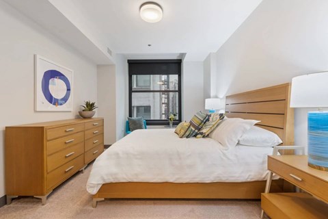 A bedroom with a large bed, a wooden dresser, and a window with a view of a cityscape.