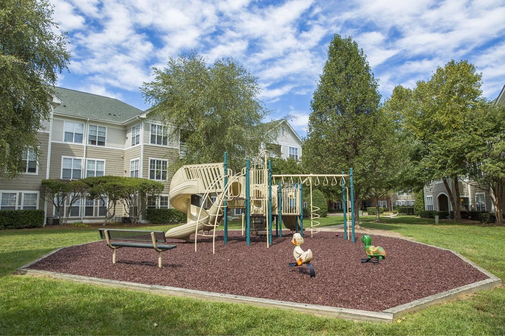 A playground with a slide, swings, and a seesaw in the foreground with apartment buildings in the background.