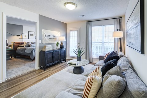 A living room with a grey sofa and a bedroom with a bed and a nightstand in the background.