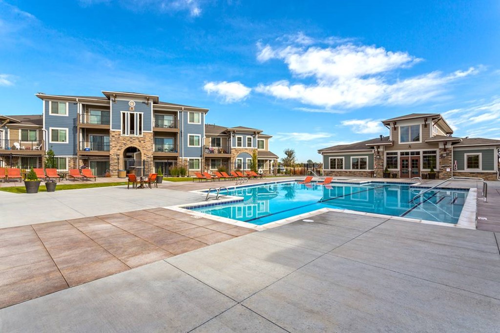 A large swimming pool is surrounded by a patio and chairs in front of apartment buildings.