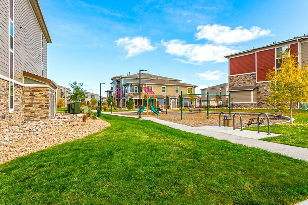 A playground with a slide and swings is surrounded by a grassy area and apartment buildings.