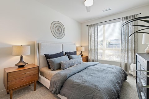 Brookside, Kansas City, MO, Apartments Near Country Club Plaza - Brookside Commons - Bedroom with Carpet Flooring, Large Bed, Nightstands, Lamps, Dresser, Wall Decor, and a Large Window with Blinds and Curtains.