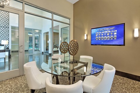 A modern dining room with a glass table and chairs, a TV displaying a 7 day forecast, and a view of a hallway through the glass doors.