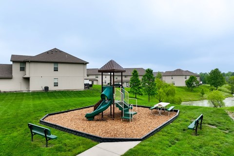 Townhomes in Peculiar, MO - Bradley’s Crossing - An Outdoor Playground Featuring a Play Structure on a Wood Fiber Surface Surrounded by Lush Grass, Trees, Benches, and Townhomes.