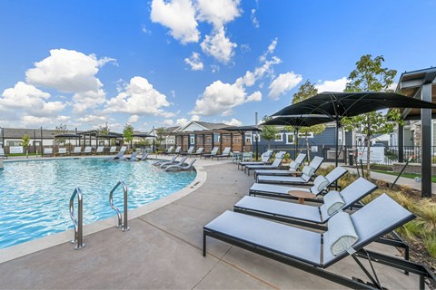 A pool with sun loungers and umbrellas is surrounded by buildings.