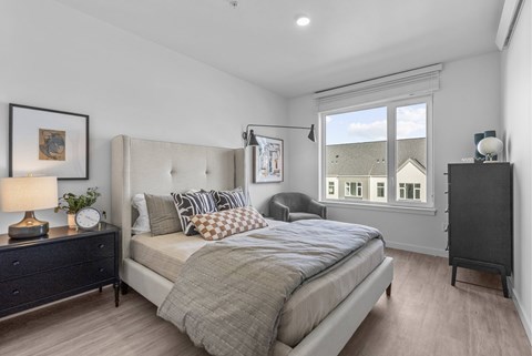 A bedroom with a large bed, a nightstand with a lamp, and a window with a view of a house.