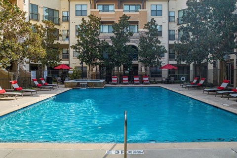 A swimming pool in front of a building with red umbrellas and lounge chairs.
