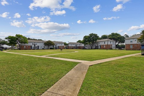 A park with a pathway in the middle of a grassy area with buildings in the background.