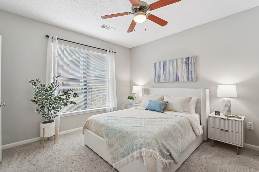 A bedroom with a large bed, a ceiling fan, and a window with a plant on the windowsill.