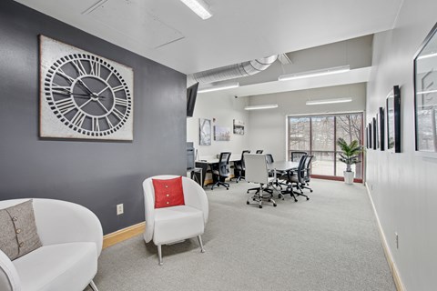 A large clock hangs on a wall above a white chair with a red pillow.