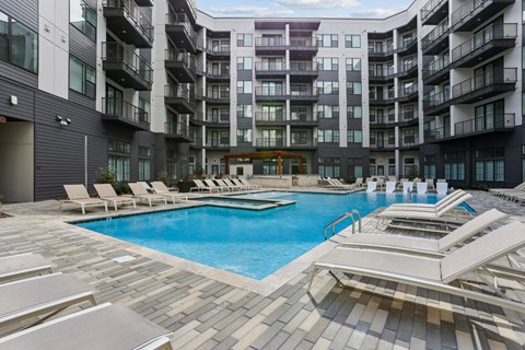 A large swimming pool surrounded by sun loungers in a courtyard of apartment buildings.