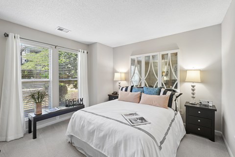 A bedroom with a large bed, a window with white curtains, and a nightstand with a lamp.