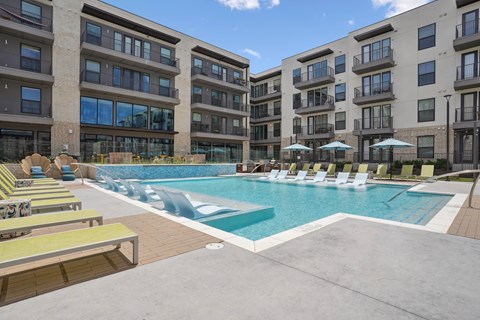 A swimming pool surrounded by sun loungers and umbrellas in front of apartment buildings.