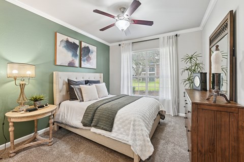 A bedroom with a bed, a nightstand, a table, a lamp, a plant, and a mirror.