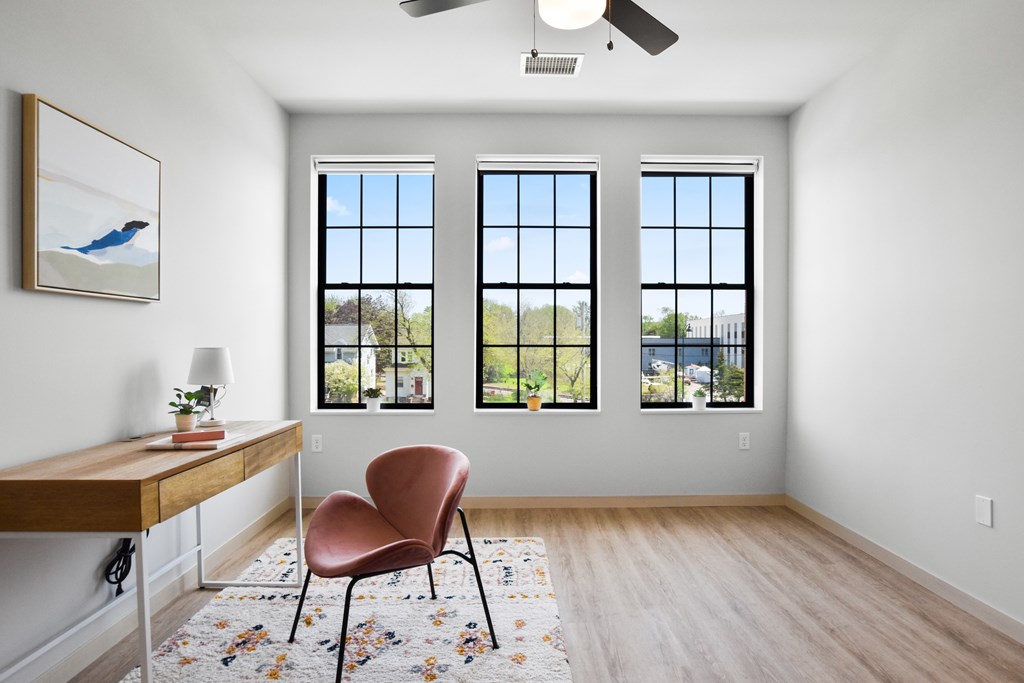 A room with a chair and a table in front of three windows.
