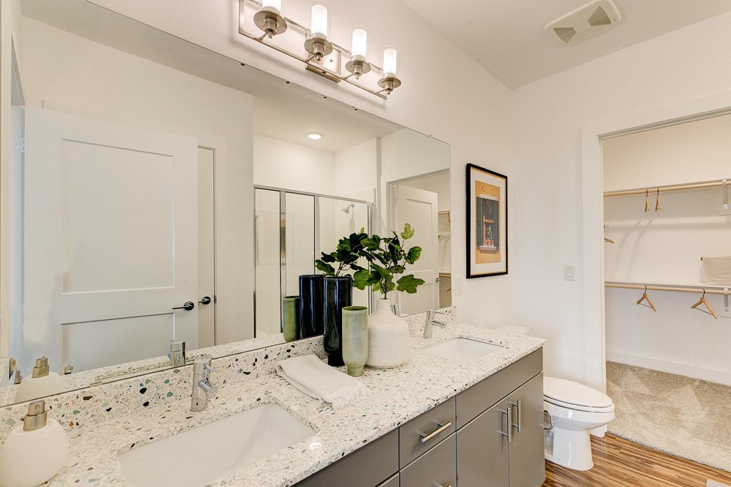 Modern bathroom here at Florence at the Harbor Apartment Homes with a large mirror, dual sinks, and terrazzo countertop. Decorative plants and towels are placed neatly. Open closet and shower visible.