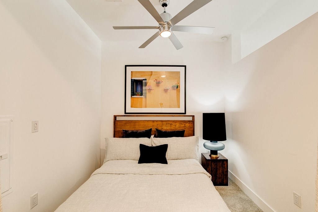 Cozy bedroom here at Florence at the Harbor Apartment Homes with a white bed, a black accent pillow, wooden headboard, and vibrant artwork above. A ceiling fan and a stylish lamp add warmth.