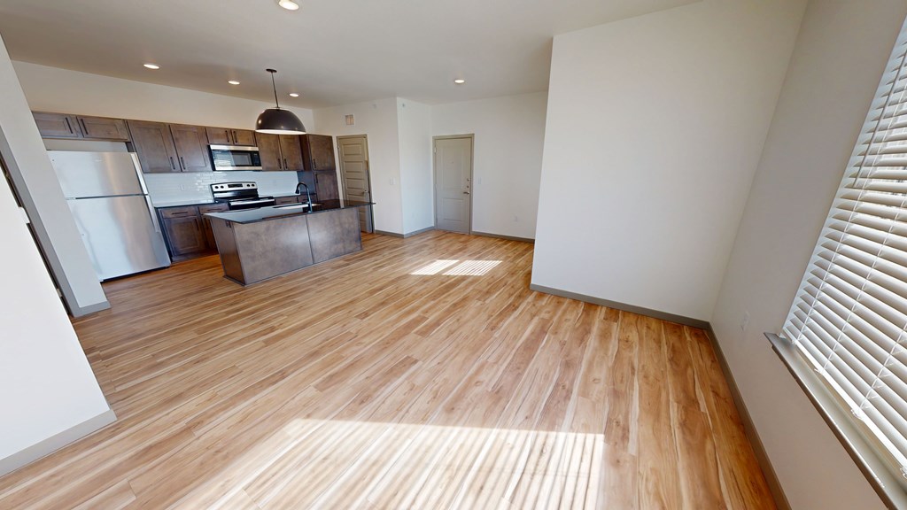 Bright open-plan kitchen and living area here at Trailwinds Apartment Homes with wood floors. Modern kitchen features stainless steel appliances and dark cabinets. Large windows allow natural light.