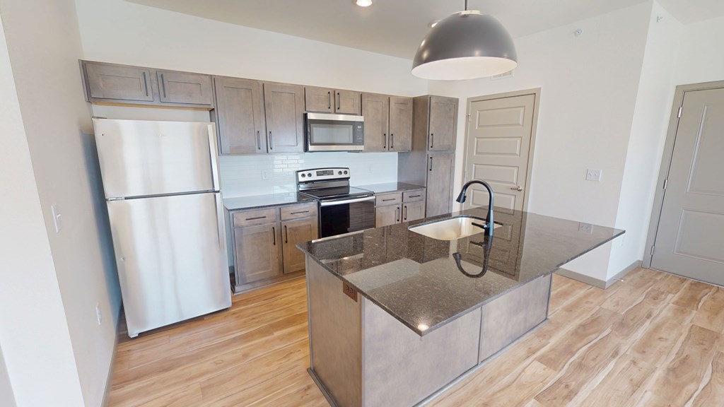 Modern kitchen here at Trailwinds Apartment Homes with wooden cabinets, stainless steel appliances, and a large island with a dark granite countertop under a sleek pendant light.