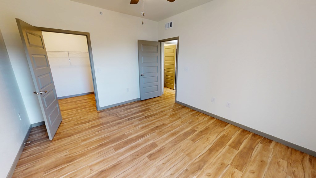 Empty bedroom here at Trailwinds Apartment Homes with hardwood floors and white walls, featuring two open gray doors leading to a closet and another room, creating a spacious, inviting feel.