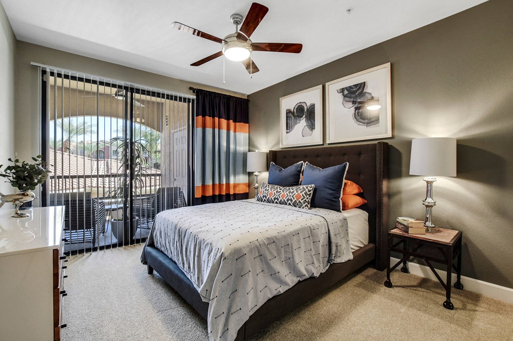 Cozy bedroom here at Desert Club Apartments with a dark upholstered bed, blue and orange pillows, abstract art, and a ceiling fan. Sliding glass door opens to a balcony.