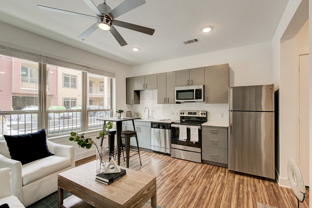 Modern living room and kitchen here at Florence at the Harbor Apartment Homes with light wood flooring, stainless steel appliances, gray cabinets, and a cozy seating area. Large windows bring in natural light.
